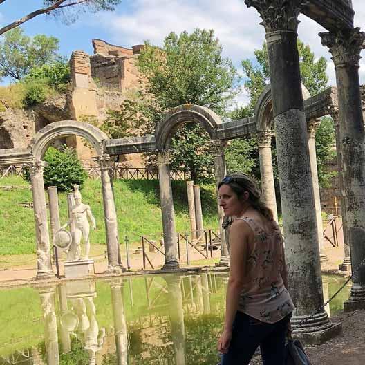 A student stands at a historical site, 在水池周围有一组拱门，背景是一尊雕像